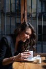 Vista laterale di bella donna con i capelli lunghi seduti al tavolo di legno dalla finestra e fare il tè — Foto stock