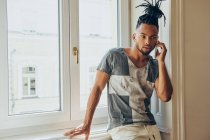 Joven hombre afroamericano con peinado creativo de pie apoyado en el alféizar de la ventana y hablando por teléfono móvil - foto de stock