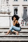 Femme ethnique en jeans et débardeur relaxant et bronzant sur des escaliers en pierre sur fond urbain — Photo de stock
