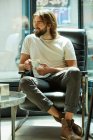 Joven hombre guapo barbudo sentado sosteniendo la taza de café en las manos y riendo - foto de stock