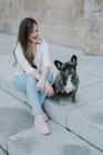 Casual young woman sitting on concrete pavement with bulldog and smiling — Stock Photo