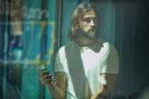 Молодой бородатый красивый мужчина опирается на стену с мобильным телефоном в руках задумчиво глядя вдоль — стоковое фото