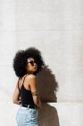Молода етнічна жінка в джинсах і танк зверху спирається на стіну і дивиться на камеру на відкритому повітрі — стокове фото