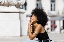 Donna etnica sorridente in jeans e canotta rilassante e prendere il sole su scale in pietra sullo sfondo urbano — Foto stock