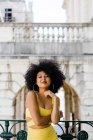 Atractiva mujer afroamericana en traje amarillo de pie y mirando a la cámara en el fondo urbano - foto de stock