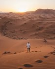 Back view of barefoot man in white summer t-shirt standing on sandy dune of endless desert in sunset, Morocco — Stock Photo