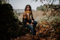 Attraktive Frau in warmer Jacke und Sonnenbrille sitzt im Herbstwald und genießt die Landschaft — Stockfoto