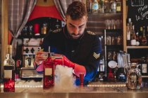 Barman in uniform and red gloves concentrated on preparing cocktail and pouring drink to smoking cocktail — Stock Photo