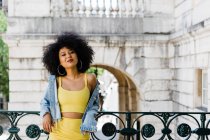 Lächelnde afrikanisch-amerikanische Frau in gelbem Anzug und Jeansjacke stehend und vor urbanem Hintergrund in die Kamera blickend — Stockfoto