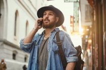 Young bearded handsome man in black hat and denim jacket merrily talking on mobile phone in street — Stock Photo