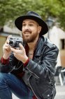 Young bearded handsome man in black hat and leather jacket taking photo in greenery street — Stock Photo