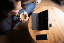 Draufsicht auf nicht wiederzuerkennende Frau, die mit Teetasse am Tisch sitzt und digitale Tablets und Smartphones nutzt — Stockfoto