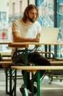 Young bearded handsome man sitting in outside cafe and working with laptop on table — Stock Photo