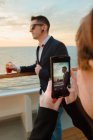 Giovane uomo bello in occhiali da sole neri con vetro di bevanda rossa in piedi sul ponte della nave, mentre la donna scattare foto sul telefono cellulare in serata di sole — Foto stock