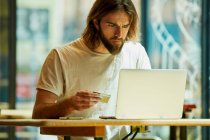 Молодий бородатий вродливий чоловік сидить у кафе і працює з ноутбуком на столі. — стокове фото