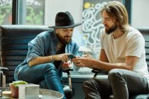 Schöner Mann mit schwarzem Hut sitzt mit Freund im Café und zeigt auf Handy — Stockfoto
