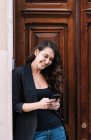 Vista lateral de la hermosa mujer usando el teléfono móvil mientras se relaja apoyado en la vieja puerta de madera - foto de stock