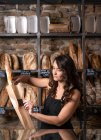 Beautiful woman in black apron putting French baguette in paper bag by metal thongs in bakery — Stock Photo