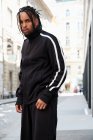 Giovane uomo etnico con i capelli intrecciati indossa tuta sportiva nera guardando la fotocamera su sfondo urbano — Foto stock