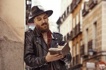 Young bearded handsome man in black hat and leather jacket with open book in hands standing near to wall in street and looking along — Stock Photo