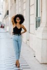 Stylish ethnic woman in jeans and tank top walking and smiling at camera outdoors — Stock Photo
