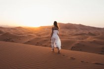 Visão traseira da mulher descalça em vestido de verão branco andando sobre duna arenosa de deserto sem fim ao pôr-do-sol, Marrocos — Fotografia de Stock
