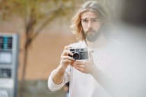 Jeune homme beau barbu en vêtements décontractés prenant des photos dans la rue — Photo de stock