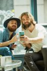 Hombre guapo en sombrero negro sentado y disfrutando del proceso de tomar selfie en el teléfono móvil con un amigo en la cafetería - foto de stock