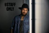 Young bearded handsome man in hat and denim shirt looking at camera in dark room — Stock Photo