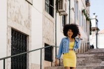 Femme afro-américaine souriante en costume jaune debout sur les escaliers et regardant la caméra sur fond urbain — Photo de stock