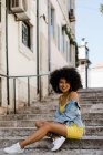 Donna afroamericana sorridente in abito giallo e giacca di jeans seduta e guardando la fotocamera su sfondo urbano — Foto stock