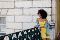 Thoughtful African American woman in yellow suit and denim jacket standing and looking away on urban background — Stock Photo