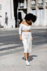Afroamerikanerin im weißen modischen Outfit steht am Straßenrand vor urbanem Hintergrund — Stockfoto
