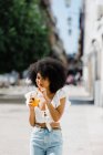 Alegre mujer afroamericana de moda bebiendo jugo de naranja y mirando a la cámara en el fondo urbano - foto de stock