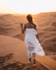 Back view of barefoot woman in white summer dress walking on sandy dune of endless desert in sunset, Morocco — Stock Photo