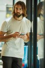 Молодой бородатый красивый мужчина, держа в руках мобильный телефон и обмениваясь сообщениями, опираясь на зеркальную поверхность — стоковое фото