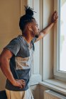 Hombre étnico serio con peinado creativo escuchar música y apoyarse en el marco de la ventana - foto de stock