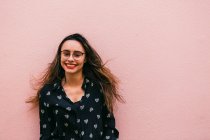 Очаровательная молодая женщина в очках и рубашке улыбается и смотрит в камеру на розовую стену — стоковое фото