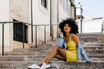 Elegante donna afroamericana in tuta gialla e denim seduta e guardando la fotocamera su sfondo urbano — Foto stock