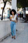 Cheerful trendy African American woman drinking orange juice and looking at camera on urban background — Stock Photo