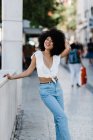 Весёлая афроамериканка в джинсах и топе расслабляется на каменных перилах и смотрит на камеру на открытом воздухе — стоковое фото