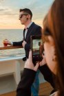 Молодий красивий чоловік в чорних сонцезахисних окулярах зі склянкою червоного напою стоїть на корабельній палубі, а жінка фотографує мобільний телефон у сонячний вечір — стокове фото