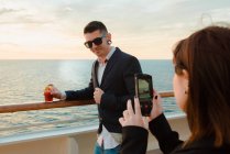 Jovem bonito homem no preto óculos de sol com vidro de bebida vermelha de pé no convés do navio, enquanto a mulher tirar foto no celular na noite ensolarada — Fotografia de Stock