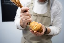 Crop man in apron and white uniform stuffing fresh croissant with chocolate cream indoors — Stock Photo
