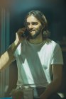 Jeune homme beau barbu gai dans un café derrière la fenêtre parlant au téléphone — Photo de stock