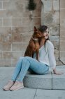 Femme décontractée avec chien de chien sur les genoux assis sur le pas de béton sur la rue et regardant loin — Photo de stock