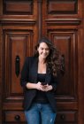 Schöne fröhliche Frau benutzt Handy, während sie sich entspannt an alte Holztür lehnt — Stockfoto