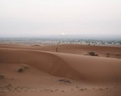 Вид на босиком женщину в летнем платье, идущую по песчаной дюне бесконечной пустыни на закате, Марокко — стоковое фото