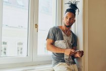 Jovem afro-americano com penteado criativo apoiado no peitoril da janela em casa usando telefone celular e ouvindo música com fones de ouvido — Fotografia de Stock