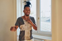 Smiling African American man with braids dancing to music with earphones at home on window background — Stock Photo